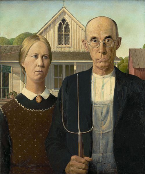 A stern man and woman stand together in front of a farmhouse, holding a pitchfork, embodying rural American life.