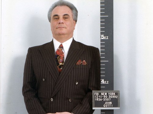 Mugshot of a man in a pinstripe suit, standing against a height chart, with a sign identifying him as John Gotti.