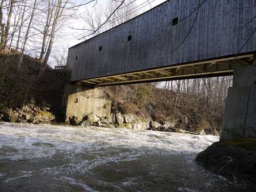 A wooden covered bridge spans a rushing river, surrounded by rocky banks and bare trees.