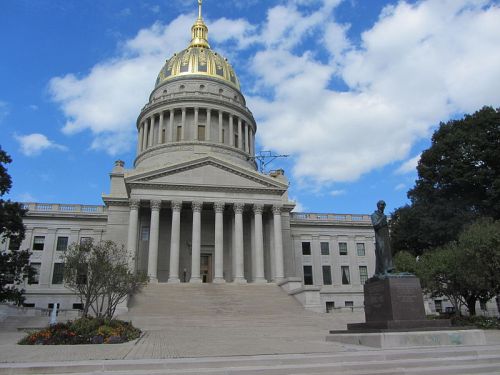 West Virginia State Capitol building with a golden dome, flanked by trees and a statue, under a blue sky with clouds.