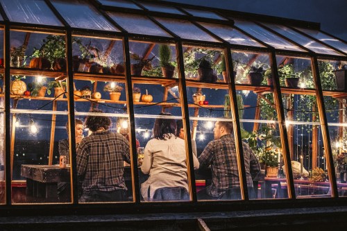 A cozy greenhouse at night, with people sitting around a table, surrounded by plants and warm lighting.