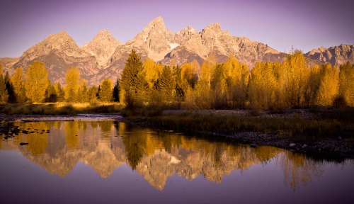 Golden autumn trees reflect in a calm river, with majestic mountains rising in the background under a purple sky.