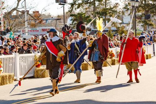 A group of historical reenactors in period costumes march in a parade, carrying spears and flags, with a crowd in the background.