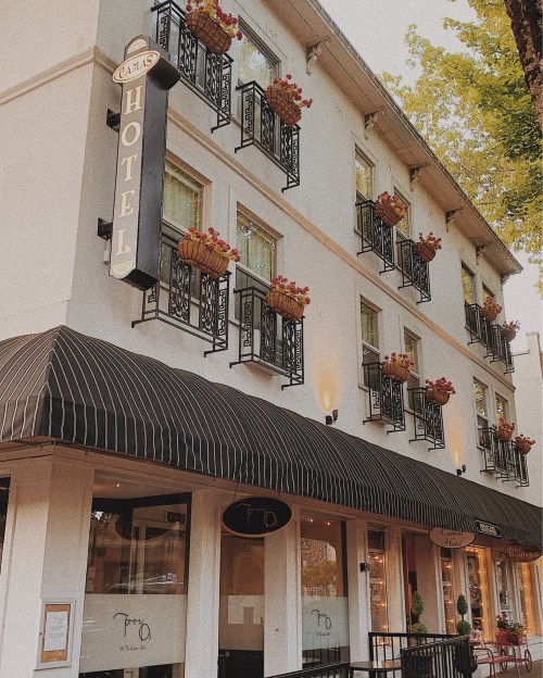A charming hotel exterior with flower-filled balconies and a striped awning, set against a tree-lined street.