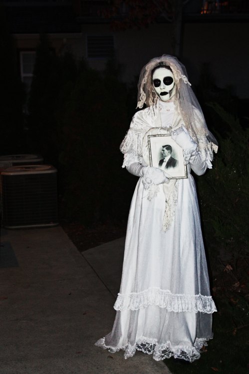 A spooky figure in a white wedding dress and veil holds a framed photo, standing in a dimly lit outdoor setting.