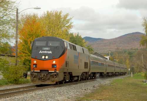 An Amtrak train travels along a track with colorful autumn trees and mountains in the background.