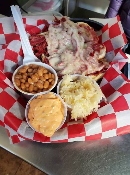 A plate with a sandwich topped with melted cheese, served with baked beans, sauerkraut, and a creamy dip.