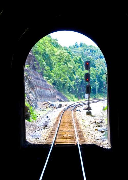 View from inside a dark tunnel, showing railroad tracks curving towards a green landscape and a signal light.