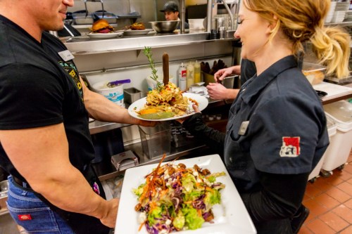 A chef hands a plate of food to a colleague in a busy kitchen, with a colorful salad on the table.