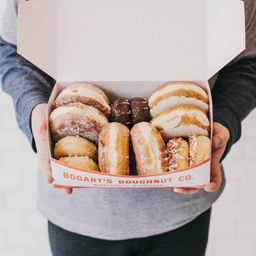 A person holding a box of assorted doughnuts from Bogart's Doughnut Co., featuring various flavors and toppings.