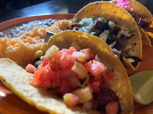 A close-up of three tacos on a plate, topped with various ingredients, alongside rice and a lime wedge.