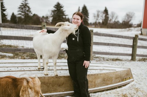A young woman in overalls smiles as a goat nuzzles her, with a dog nearby on a snowy farm.