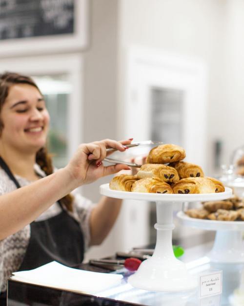 A smiling woman serves freshly baked croissants from a display stand in a bakery.
