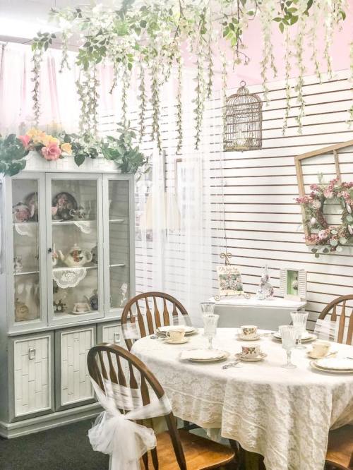 A charming dining area with a lace tablecloth, vintage dishes, and greenery hanging from the ceiling.