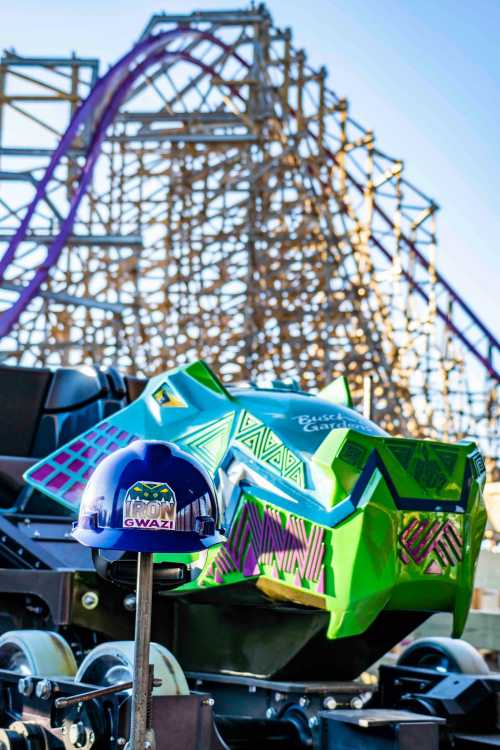 A colorful roller coaster car with a blue helmet in front, set against a wooden roller coaster structure in the background.