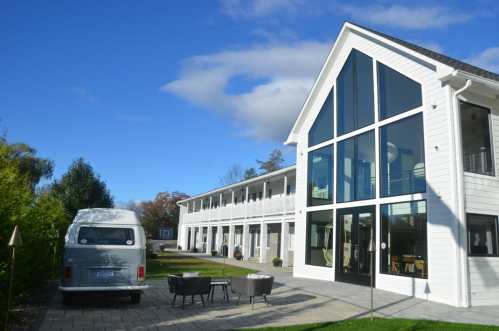 A modern white building with large windows, surrounded by greenery and a van parked nearby under a blue sky.