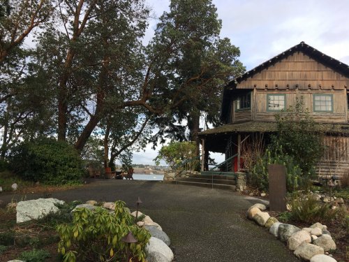 A rustic wooden house surrounded by trees, with a pathway leading to the entrance and a view of the water in the background.