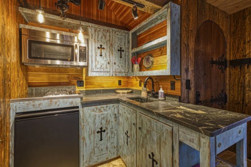 Rustic kitchen with blue cabinets featuring cross designs, a microwave, and dark countertops in a wooden setting.