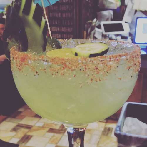A vibrant margarita in a large glass, garnished with a lime slice, cucumber, and a colorful umbrella.