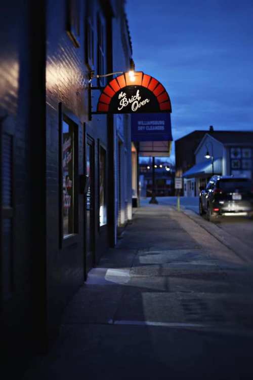 A dimly lit street scene at dusk featuring a sign for "The Brick Oven" restaurant.