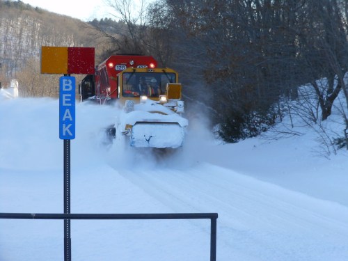 A snowplow clears a snowy road, with trees in the background and a sign labeled "BEAK" nearby.