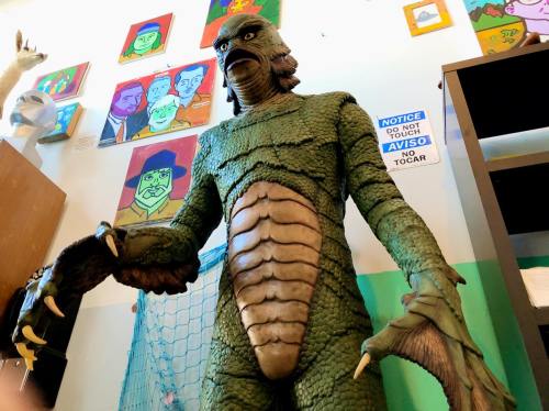 A life-sized creature figure with green scales and claws stands in a room decorated with colorful artwork.