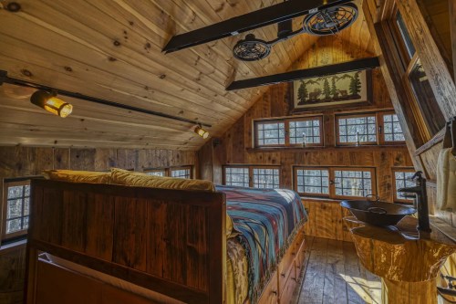 Cozy wooden bedroom with a sloped ceiling, large windows, and rustic decor, featuring a bed and unique sink.