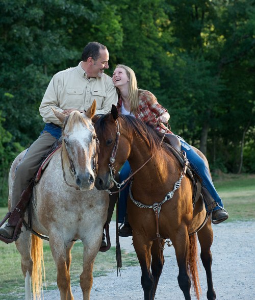 A man and woman share a joyful moment on horseback, smiling at each other in a natural outdoor setting.