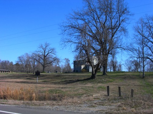 A rural scene featuring a house surrounded by bare trees and a clear blue sky, with a grassy area in the foreground.