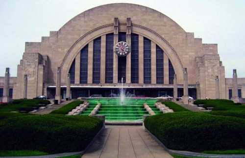 Historic train station with a large clock, fountain, and landscaped gardens in front. Overcast sky above.