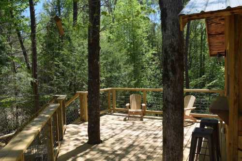 A wooden deck surrounded by lush green trees, featuring two chairs and a rustic cabin structure nearby.