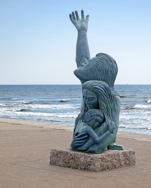 A bronze sculpture of a woman embracing a child, with another hand raised, set against a beach and ocean backdrop.