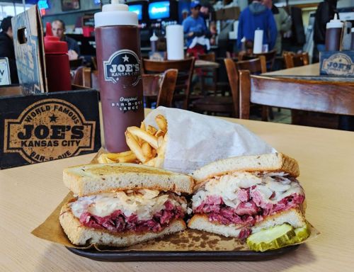 A hearty sandwich with meat and toppings, served with fries and a bottle of barbecue sauce in a casual dining setting.