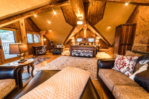 Cozy attic bedroom with wooden beams, a large bed, elegant decor, and comfortable seating areas. Warm lighting enhances ambiance.