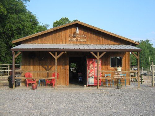 A wooden stable with a sign reading "Wranglers Riding Stables," featuring a porch and red chairs outside.