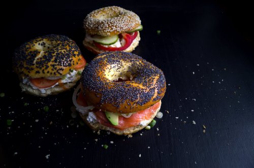 Three bagel sandwiches with various toppings, including tomatoes, cucumbers, and pickles, on a dark surface.