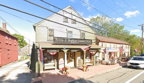 A quaint country store with a sign reading "Brown & Hopkins," featuring flags and outdoor seating.