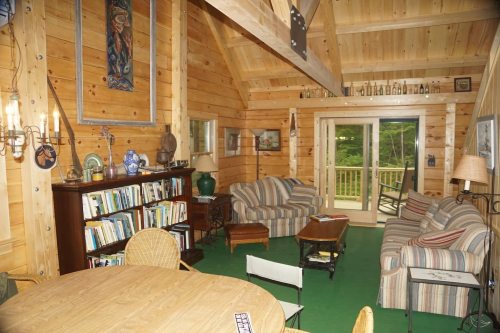 Cozy wooden cabin interior with a bookshelf, seating area, and large windows overlooking greenery.