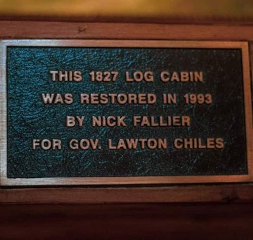 Plaque on a wooden surface reads: "This 1827 log cabin was restored in 1993 by Nick Fallier for Gov. Lawton Chiles."