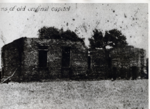 Black and white photo of the ruins of an old building, labeled as the original capitol, surrounded by trees.