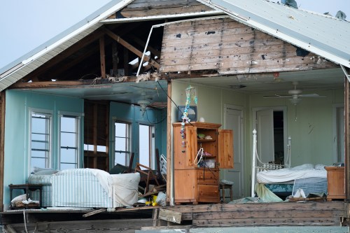 A damaged house with exposed interior, showing broken furniture and debris after a storm or disaster.
