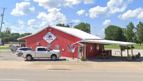 A red building with a white roof, featuring a sign, surrounded by trees and parked cars on a sunny day.
