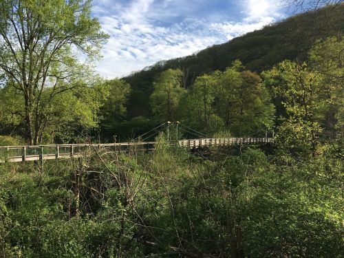 A wooden suspension bridge surrounded by lush green trees and hills under a partly cloudy sky.