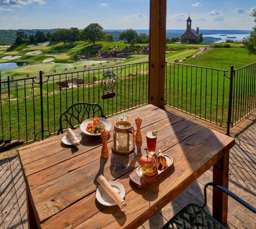 A scenic outdoor dining setup with a wooden table, plates of food, and a view of a golf course and lake.
