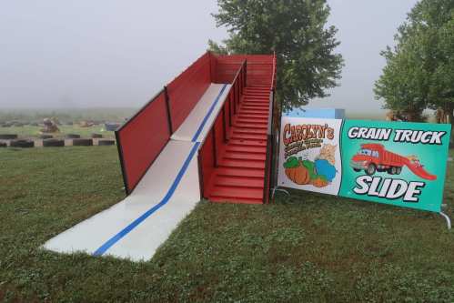 A red slide with stairs beside a sign for a grain truck slide, set in a foggy outdoor area with grass.