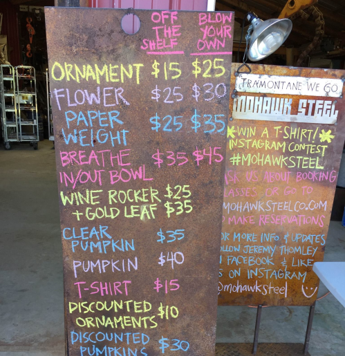 A rustic sign listing items for sale: ornaments, flower, paperweight, wine rocker, pumpkin, and T-shirts with prices.
