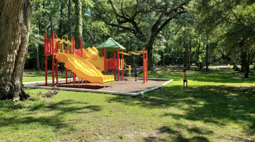 A sunny playground with a yellow slide and red structures, surrounded by trees and children playing on the grass.