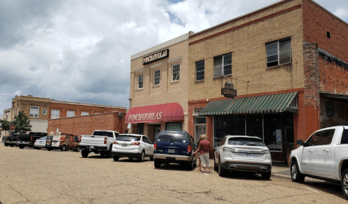 A street view of a small town with brick buildings, parked cars, and a cloudy sky.