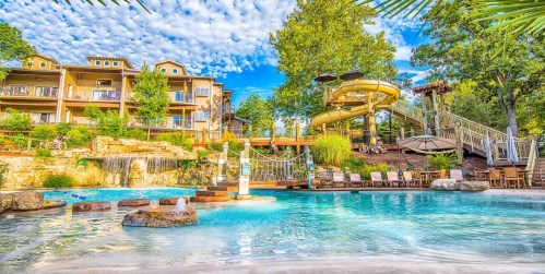 A vibrant resort pool area featuring a water slide, lush greenery, and a backdrop of charming buildings under a blue sky.