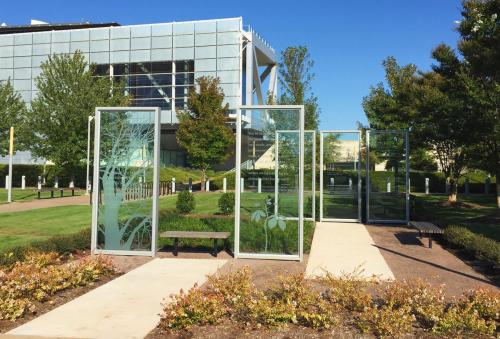 A glass structure with etched tree designs, surrounded by greenery and a pathway, near a modern building.